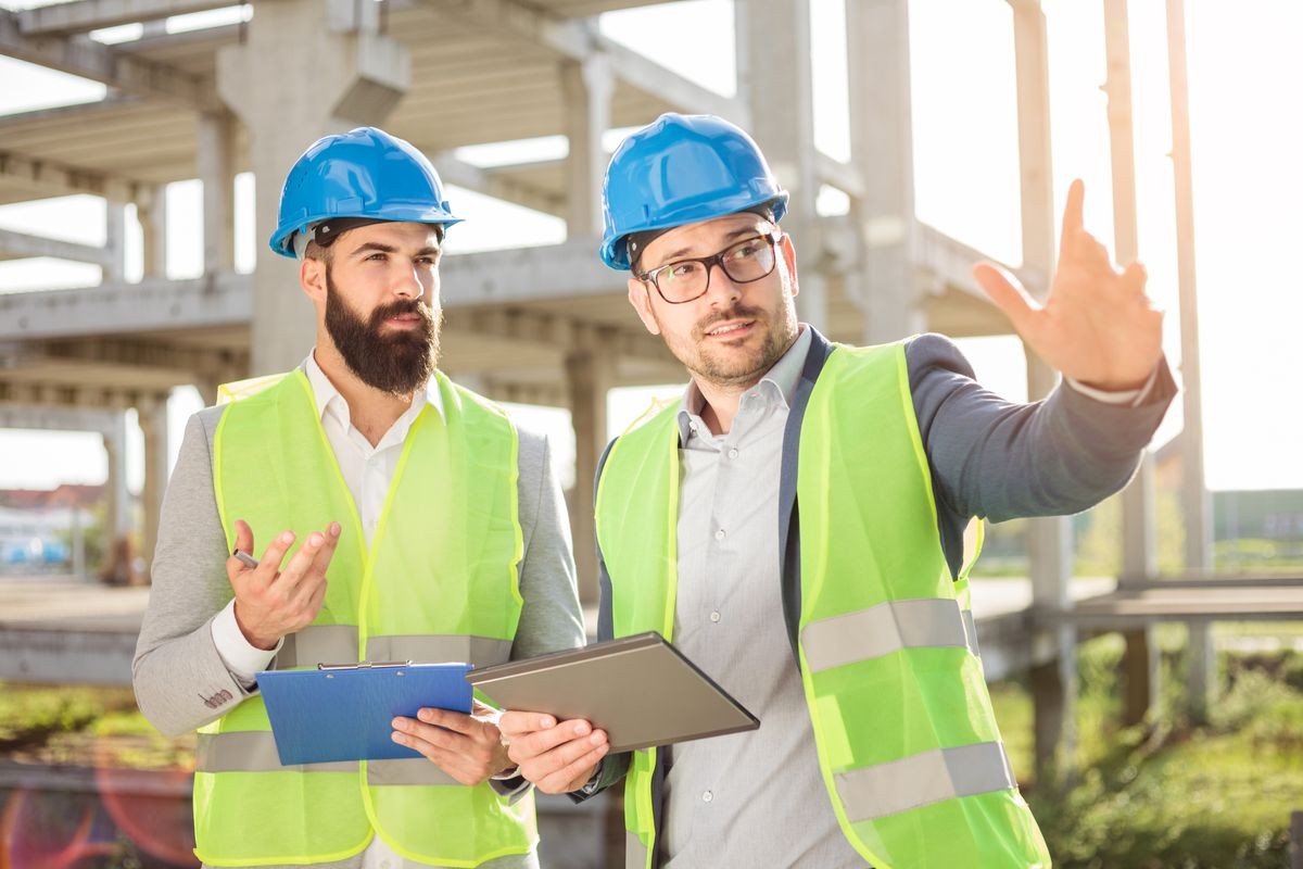 Two young male architects or business partners talking on a construction site. One man is pointing into distance with his arm, showing work progress.